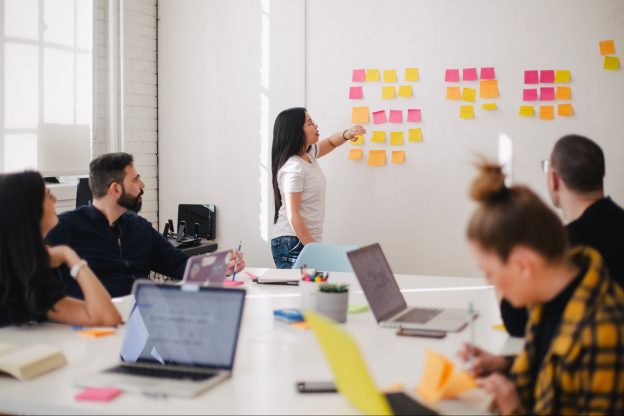 Woman placing sticking notes on a wall in front of other people sitting at a desk with their laptops